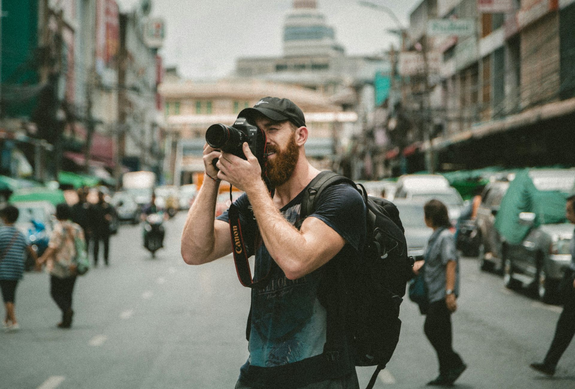 Un homme barbu avec un sac à dos prend une photo dans une rue animée.